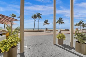 A paved walkway with palm trees and potted plants, leading to a beach area with a clear sky. - Hotel Sheldon (Hollywood)
