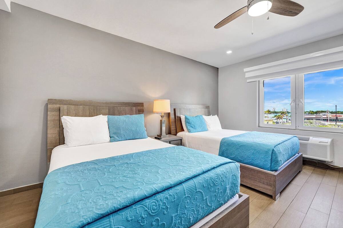 A hotel room with two beds, a ceiling fan, a nightstand with a lamp, and a large window offering a view of the city.