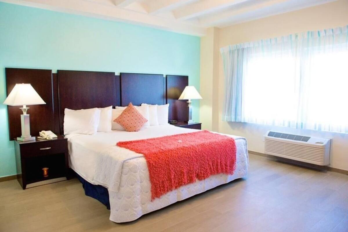 A hotel room with a bed, bedside lamps, a heater, and a window with curtains.