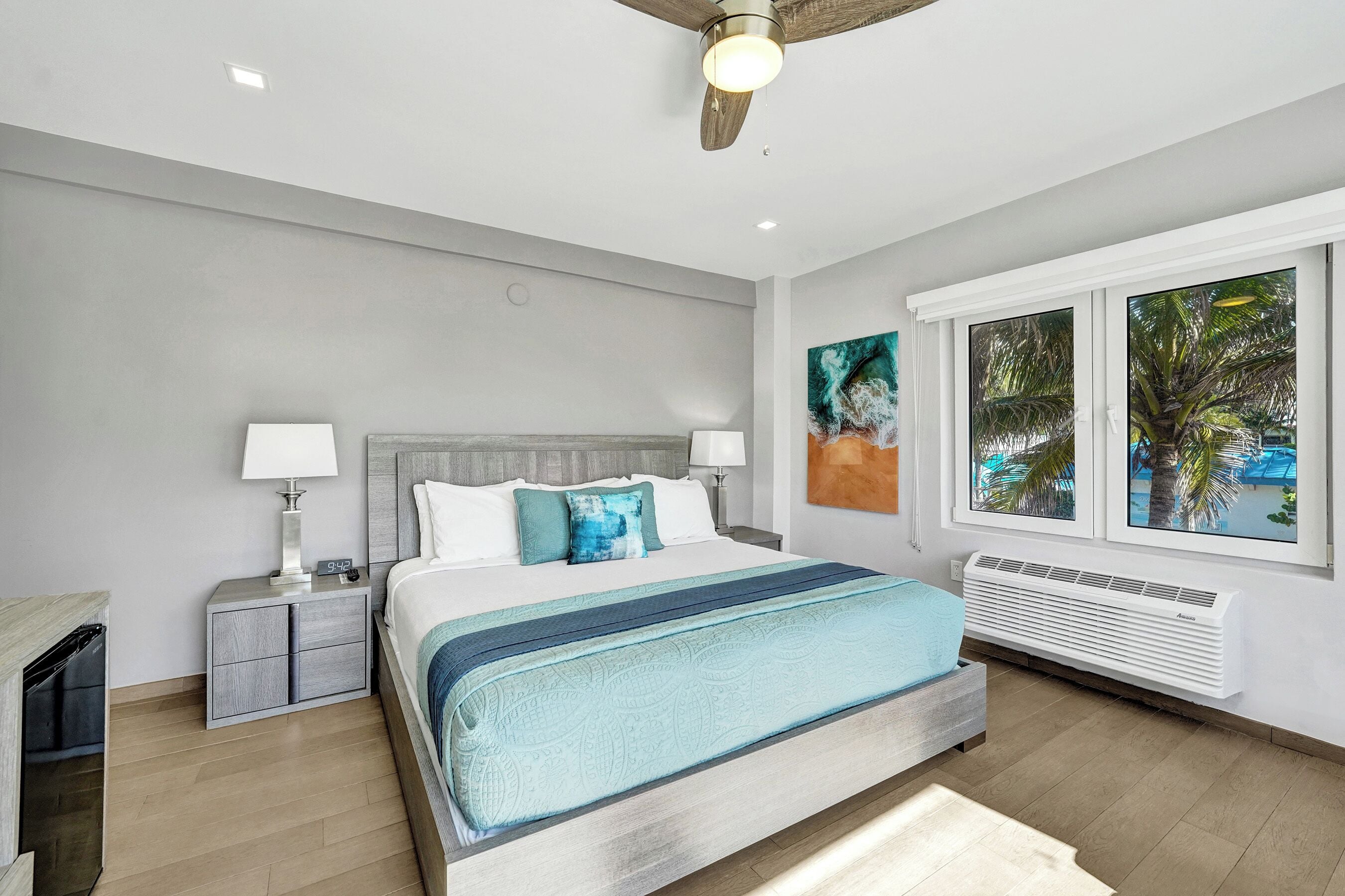 A modern bedroom with a bed, bedside tables, a ceiling fan, and a window with a view of palm trees.