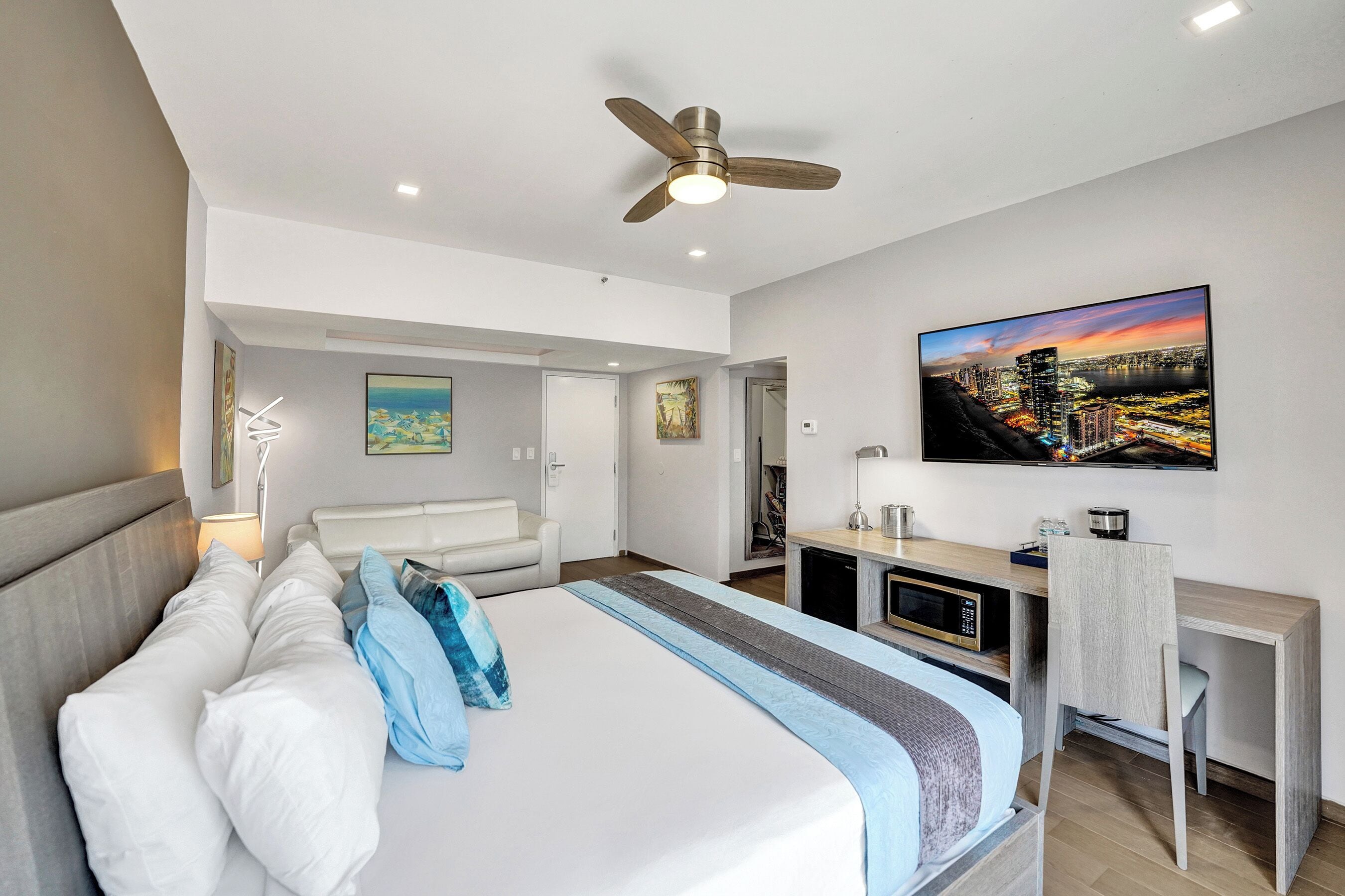 A modern hotel room with a large bed, a desk, a chair, a ceiling fan, and a cityscape painting on the wall.