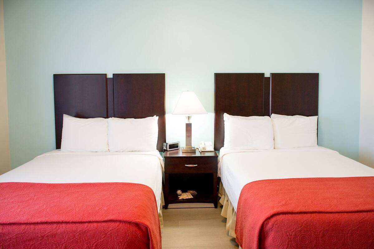 A hotel room with two beds, a nightstand, a lamp, and a small wooden chest.