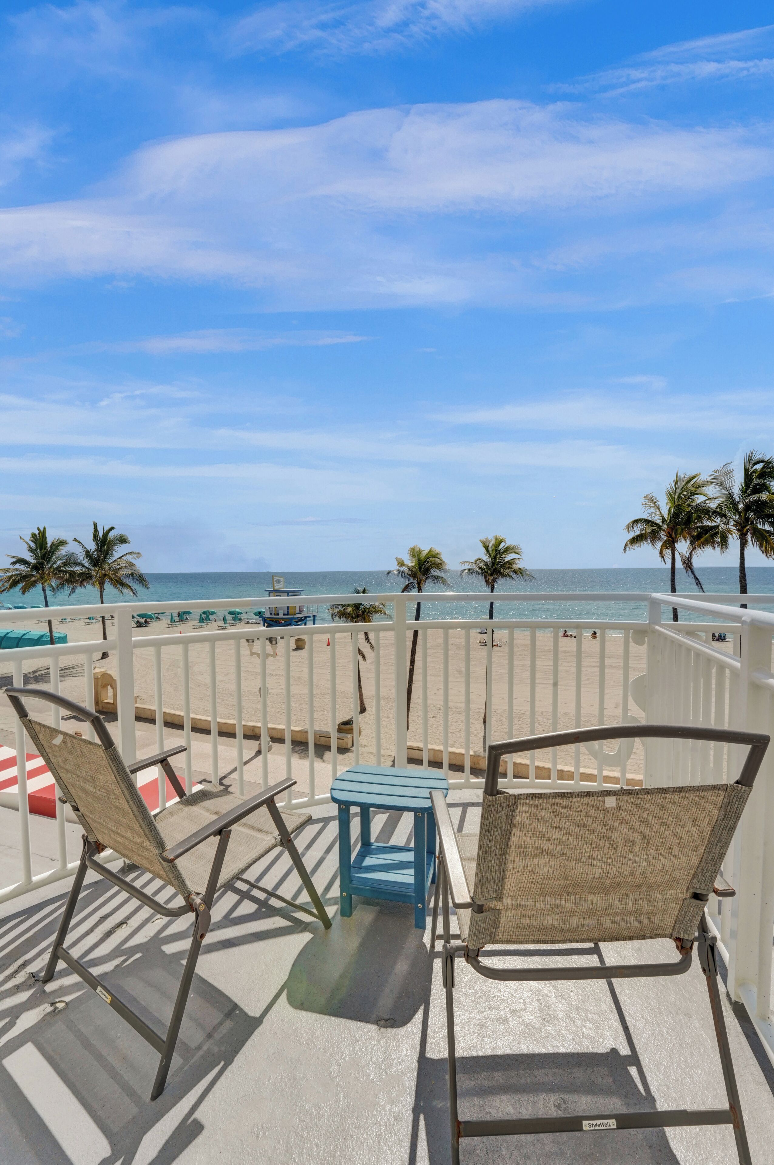 A balcony with two chairs and a small table, overlooking a beach with palm trees and the ocean.