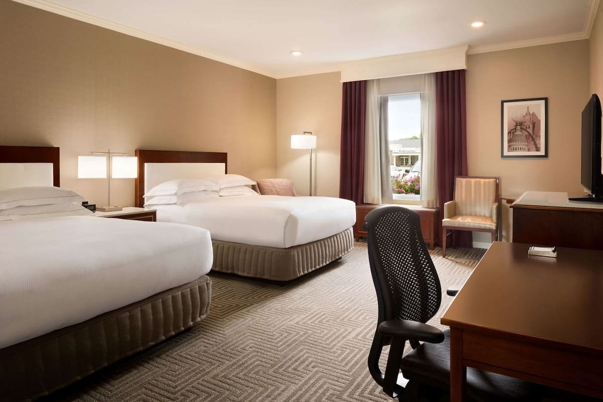 A hotel room with two beds, a desk, a chair, a TV, and a window with curtains.