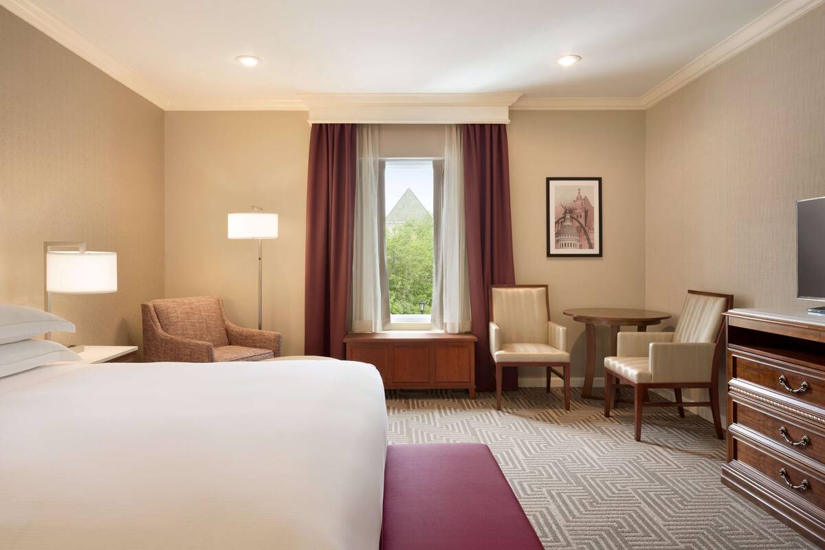 A hotel room with a large bed, two armchairs, a desk, a television, and a window with curtains.