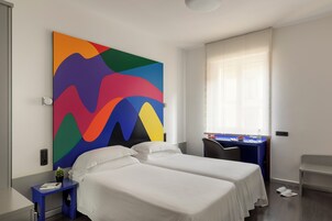Een hotelkamer met twee bedden, een bureau, een stoel, een kleurrijk abstract schilderij en een raam met zonwering.