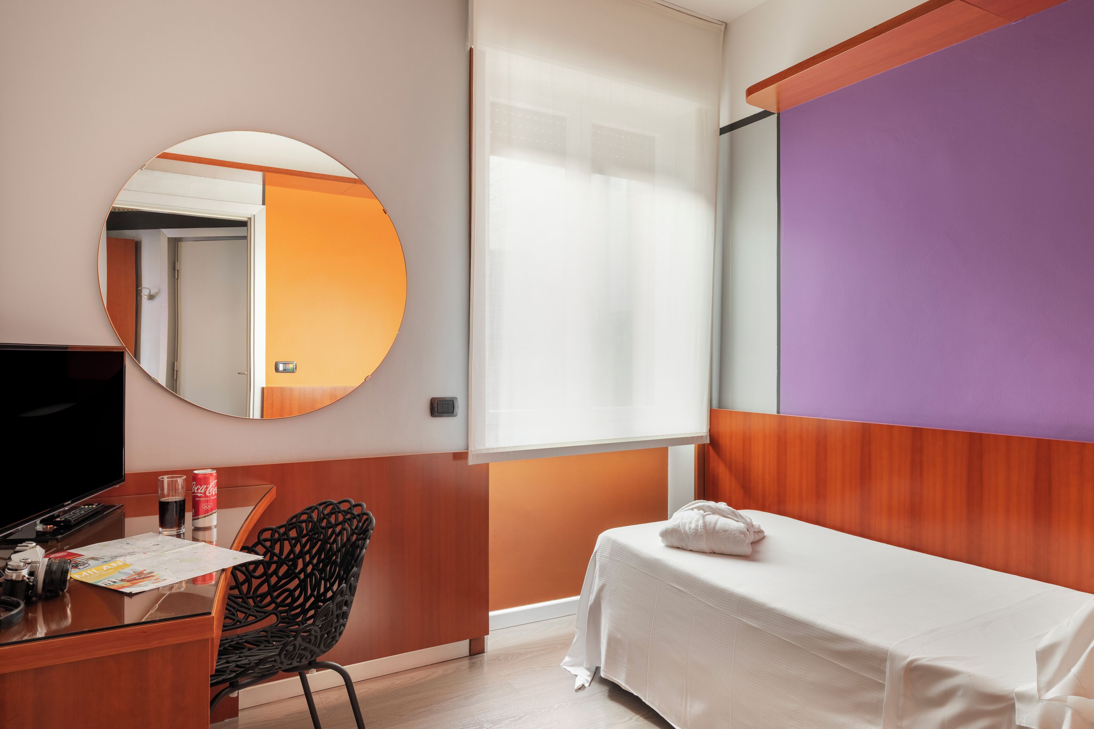 Une chambre d’hôtel comprenant un lit, un bureau avec une télévision, une chaise, un miroir rond et une fenêtre avec des stores.