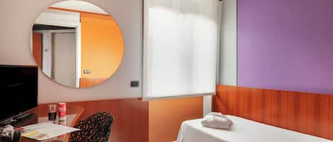Habitación de hotel con cama, escritorio con televisor, silla, espejo redondo y ventana con persianas.