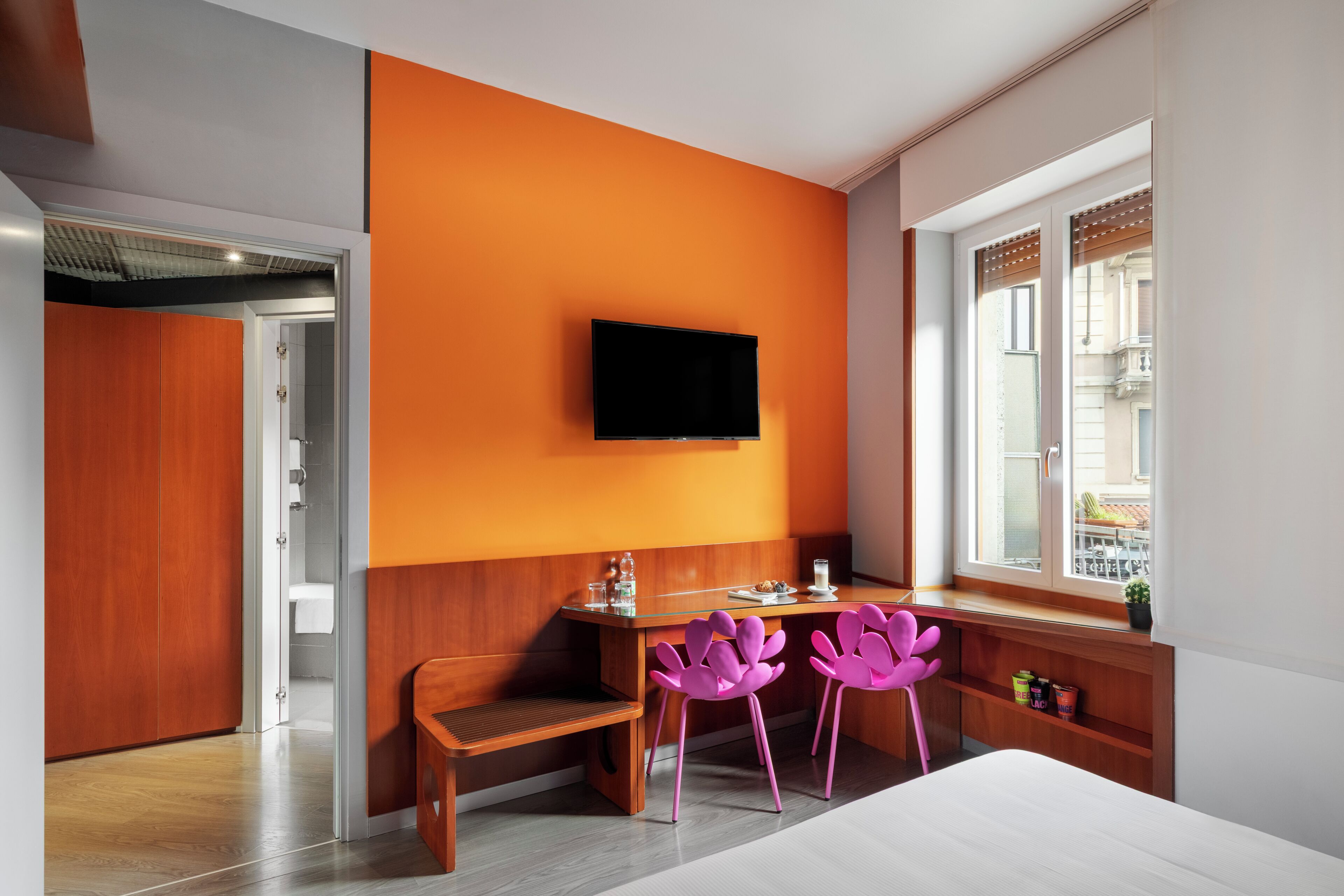 Habitación de hotel con un escritorio de madera, dos sillas rosas, un televisor de pantalla plana montado en la pared y un ventanal con vistas a edificios.