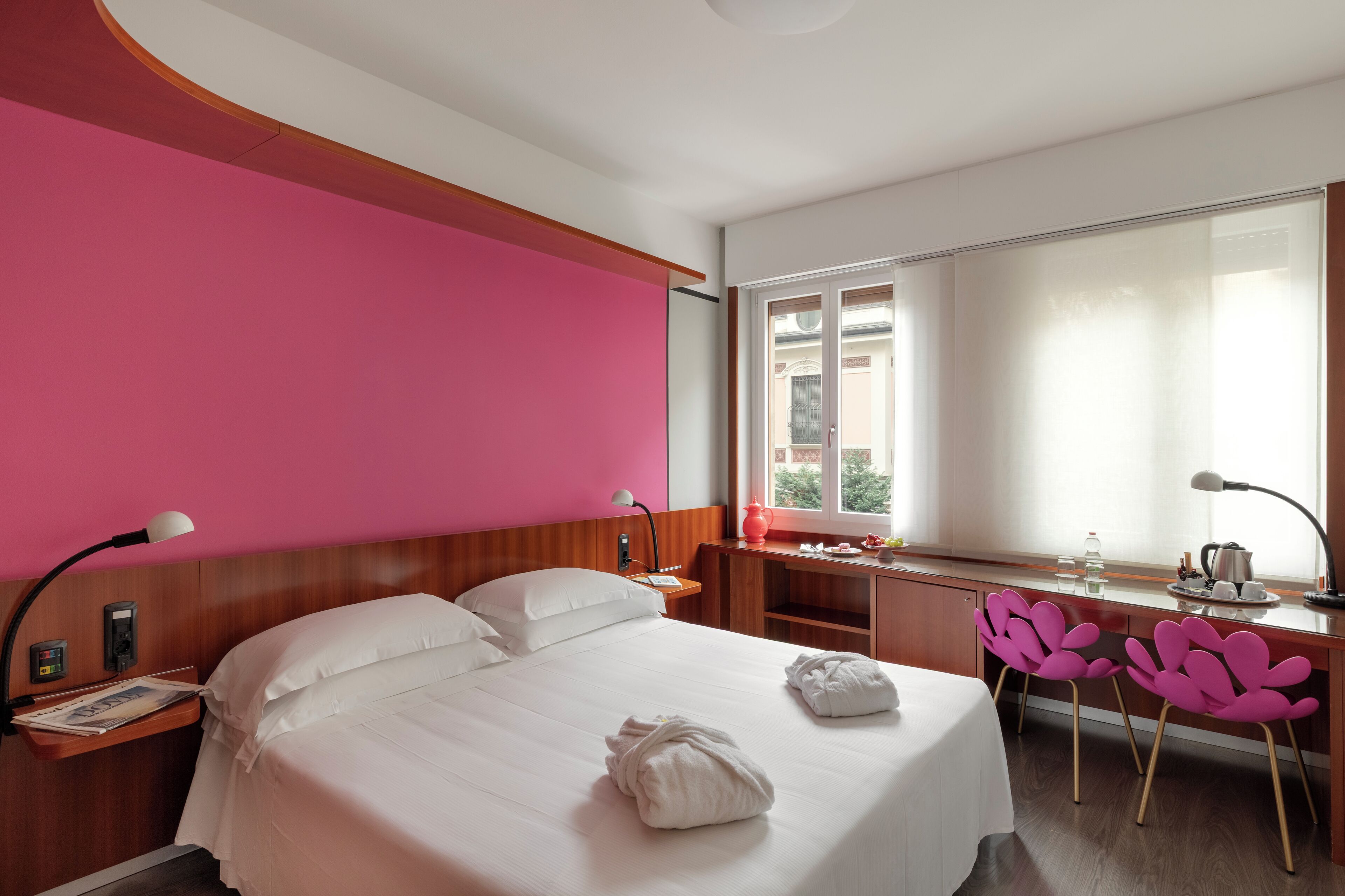 A hotel room with a bed, bedside table, lamp, desk, chair, and a window with a view.