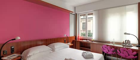 A hotel room with a bed, bedside table, lamp, desk, chair, and a window with a view.