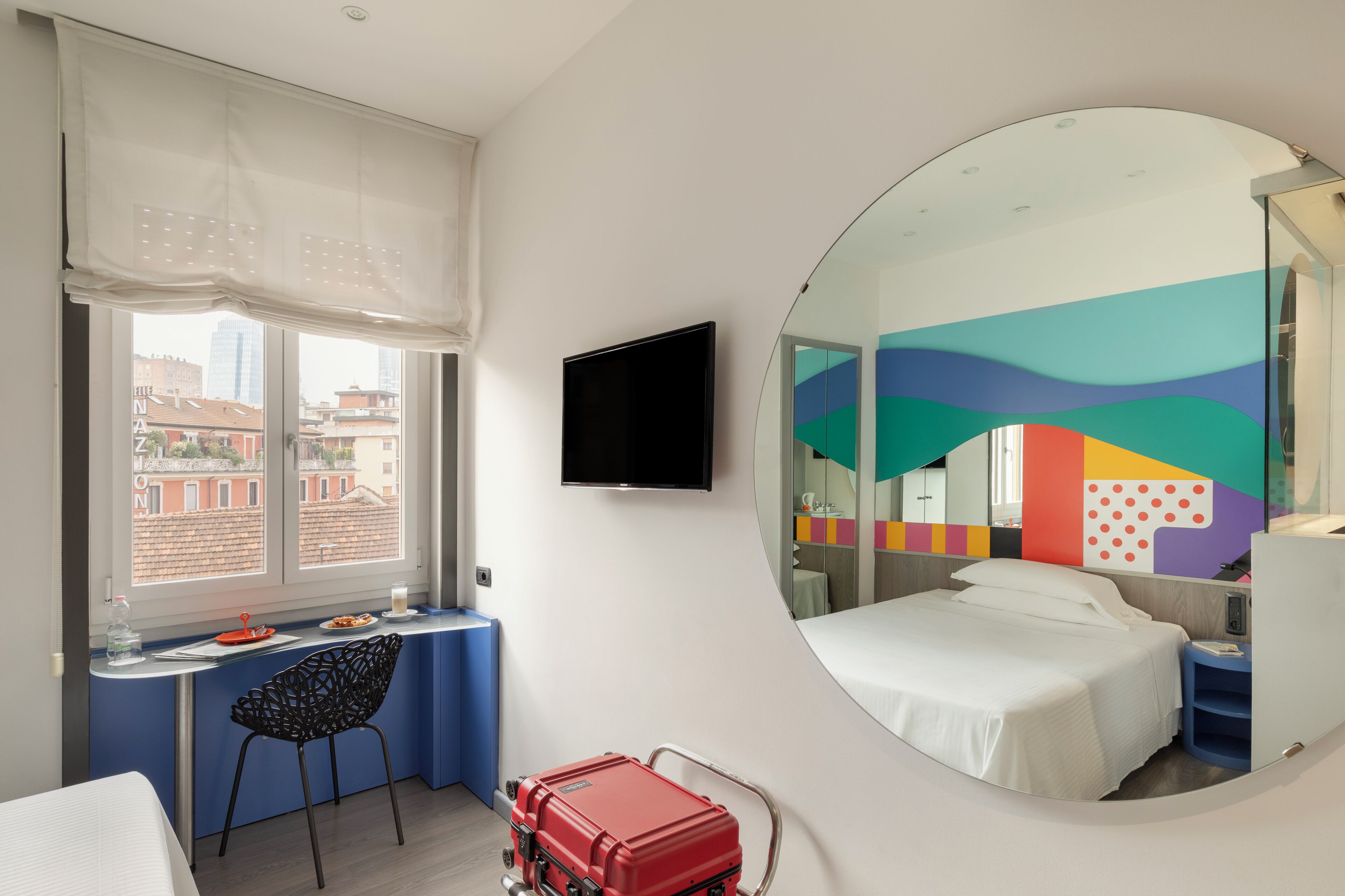 A hotel room with a bed, a desk, a chair, a TV, a suitcase, and a large mirror.