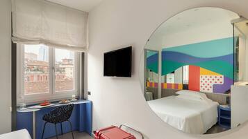 A hotel room with a bed, a desk, a chair, a TV, a suitcase, and a large mirror.