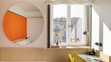 Ein modernes Hotelzimmer mit einem runden Spiegel, einem Bett, einem Schreibtisch mit Lampe und Blick auf Gebäude durch ein Fenster.