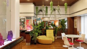 A modern interior with a wooden ceiling, hanging plants, a yellow sofa, white chairs, and a glass table. - Hotel Mediolanum (Milan)