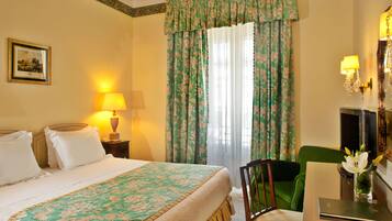 Una habitación de hotel con cama, escritorio, silla, lámpara, un cuadro en la pared y ventana con cortinas.