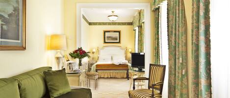 A hotel room with a green sofa, a wooden chair, a bed with a white headboard, a television, and a painting on the wall.