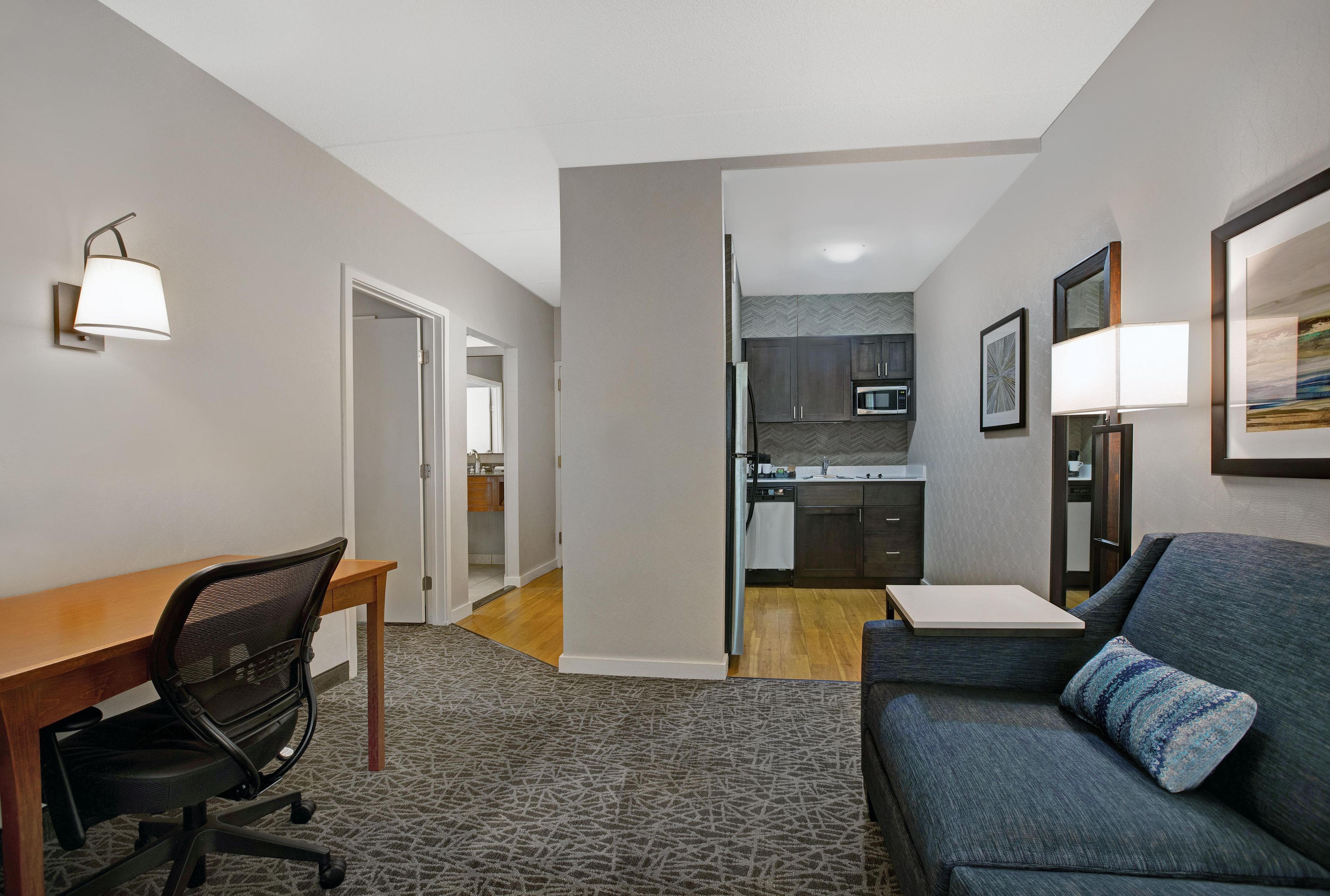 A modern living room with a sofa, a desk, and a chair. There is a kitchen area with cabinets and a microwave.