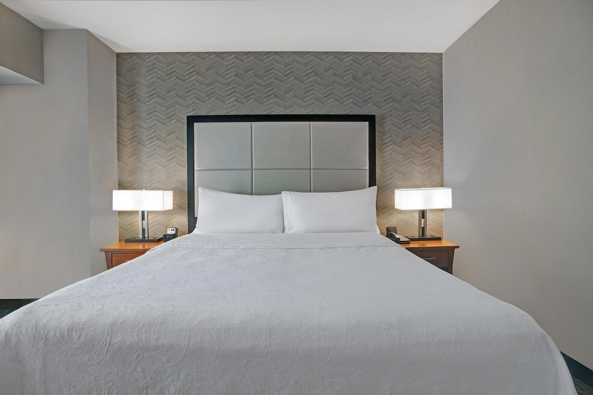 Quarto de hotel com uma cama grande, duas mesas de cabeceira com luminárias e um papel de parede em padrão losango.