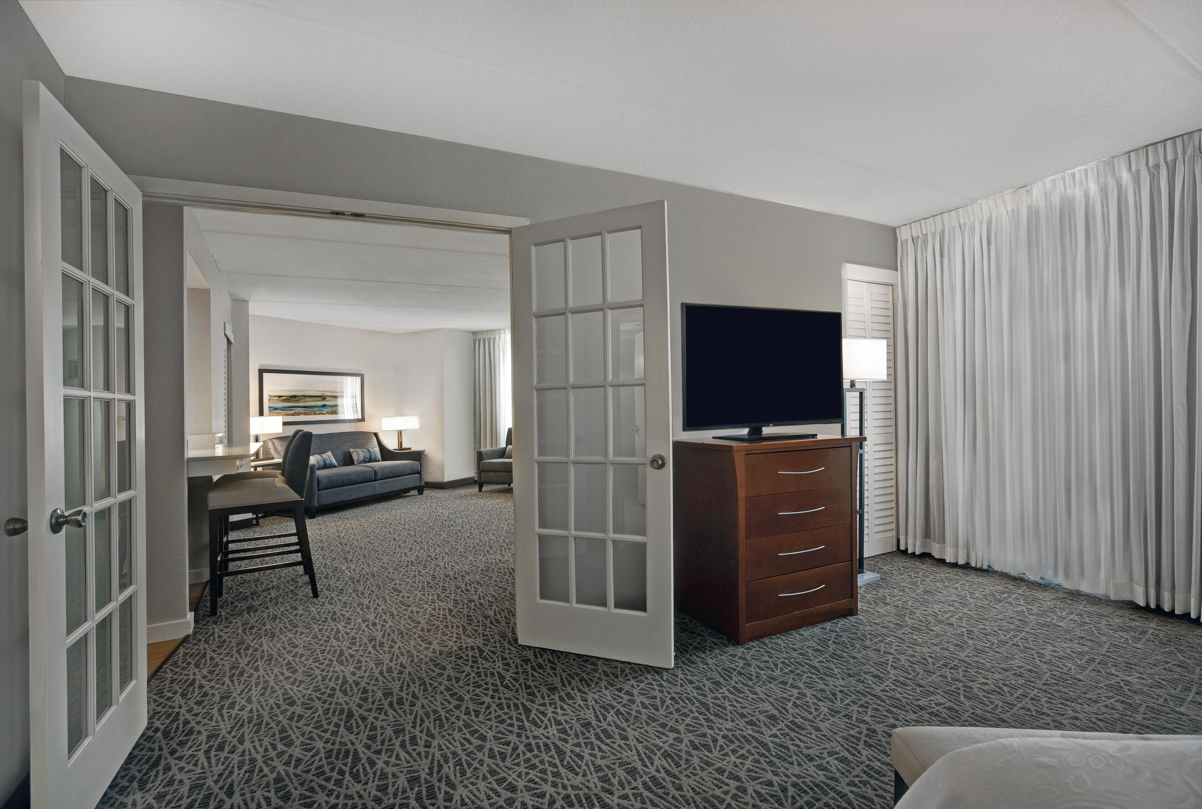 A hotel room with a large bed, a TV on a wooden dresser, a desk with a chair, and a large window with curtains.