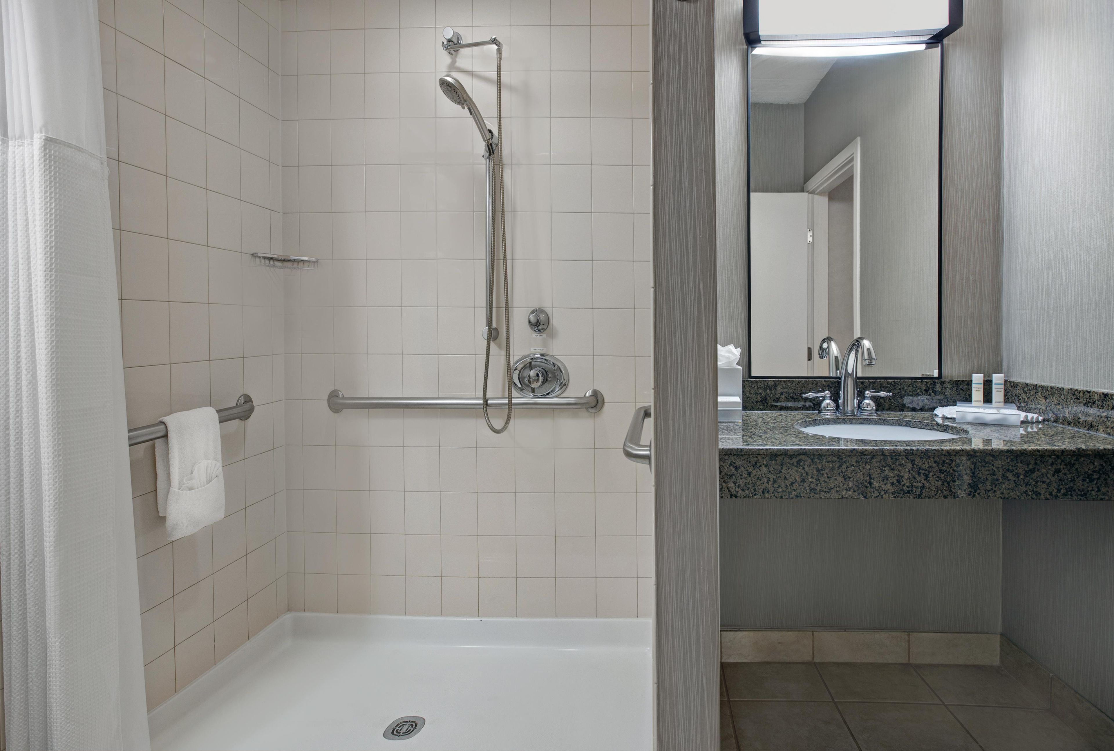 A modern bathroom with a walk-in shower, grab bars, a bathtub, and a granite countertop with a sink and mirror.