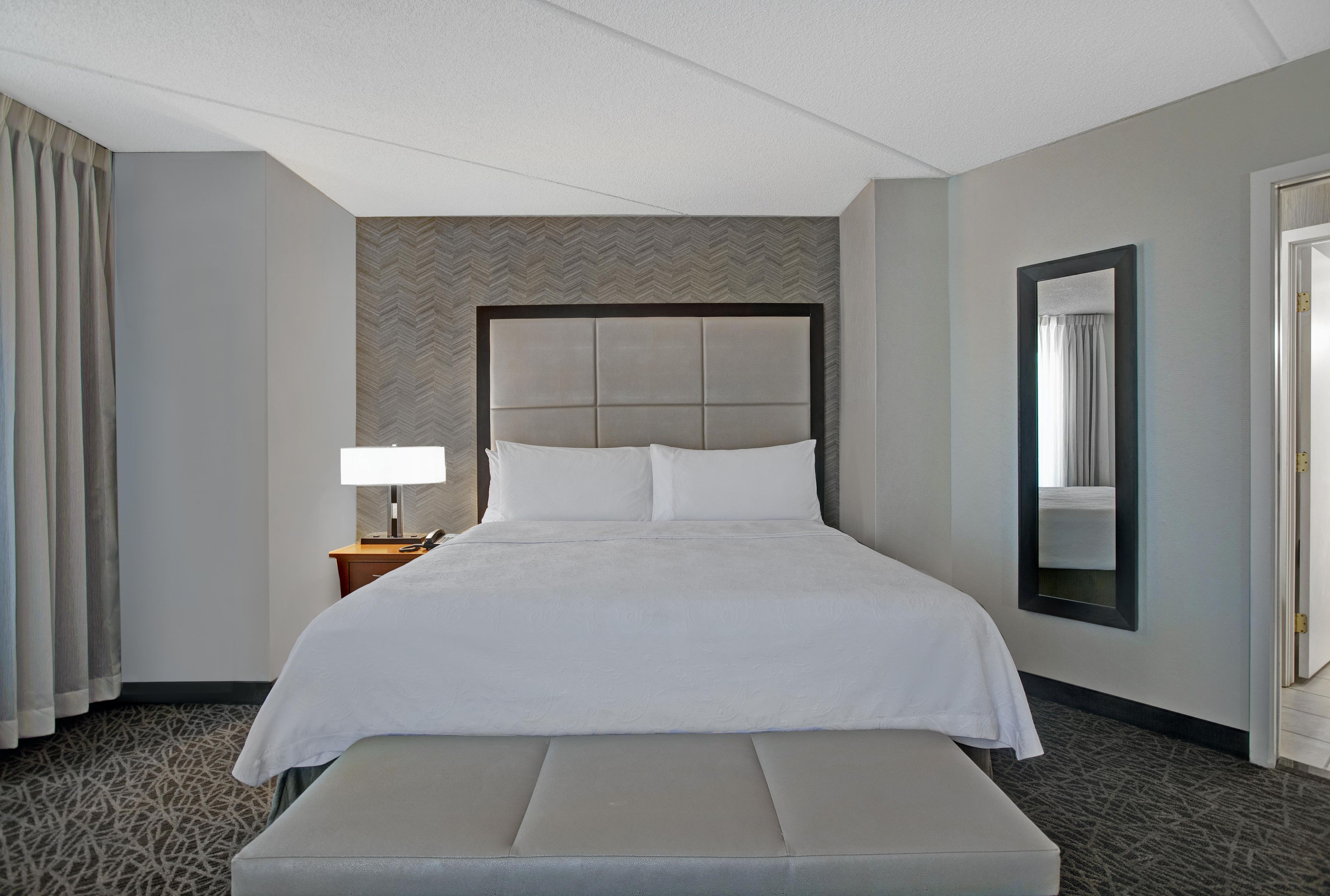 Una habitación de hotel con una cama grande, una mesita de noche con lámpara, un banco, un espejo y una alfombra estampada.