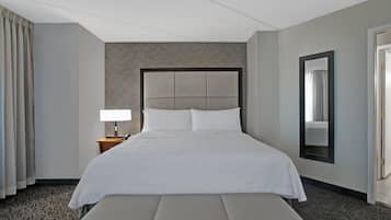 Una habitación de hotel con una cama grande, una mesita de noche con lámpara, un banco, un espejo y una alfombra estampada.