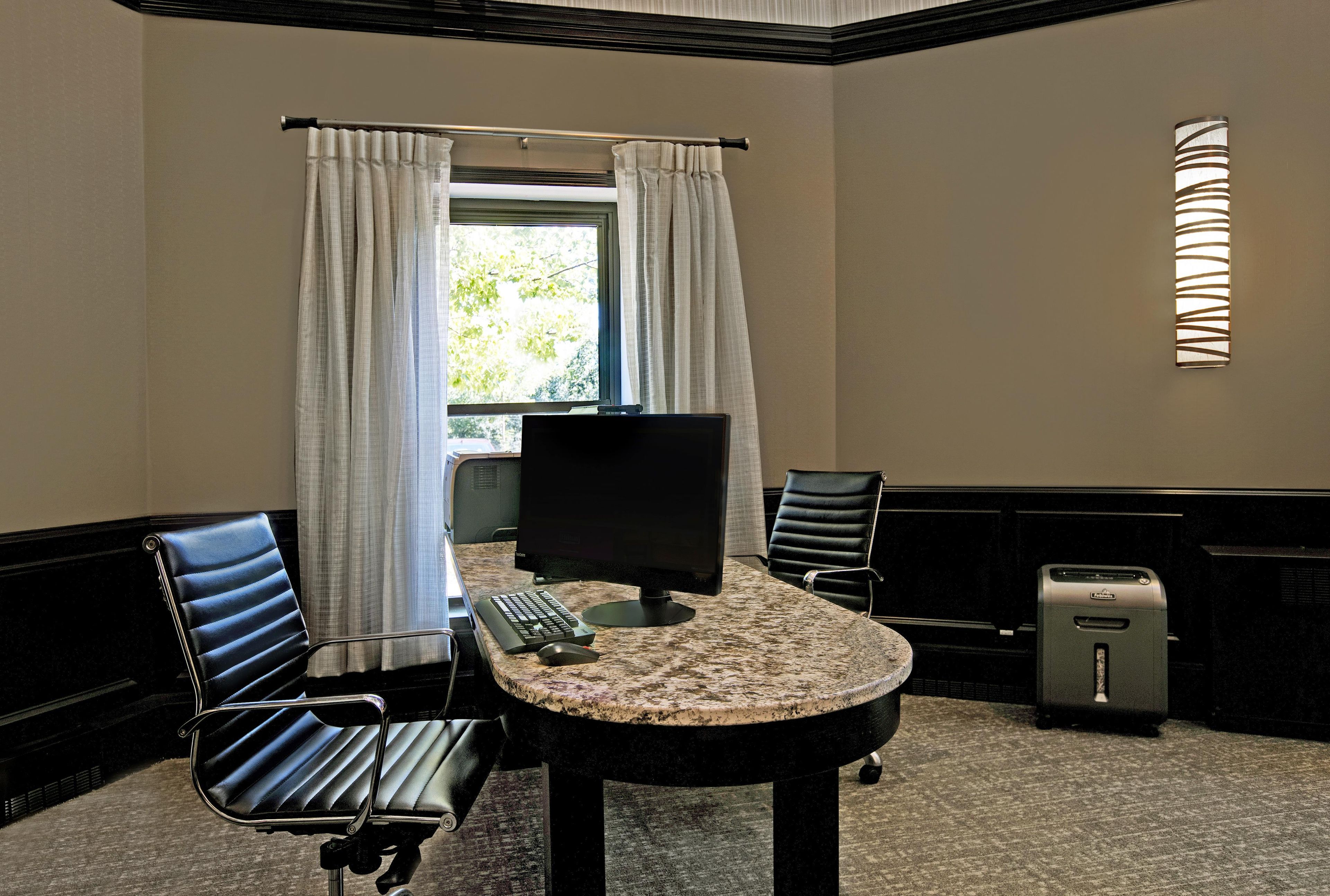 A hotel room with a desk, two chairs, a computer, a telephone, and a window with curtains.