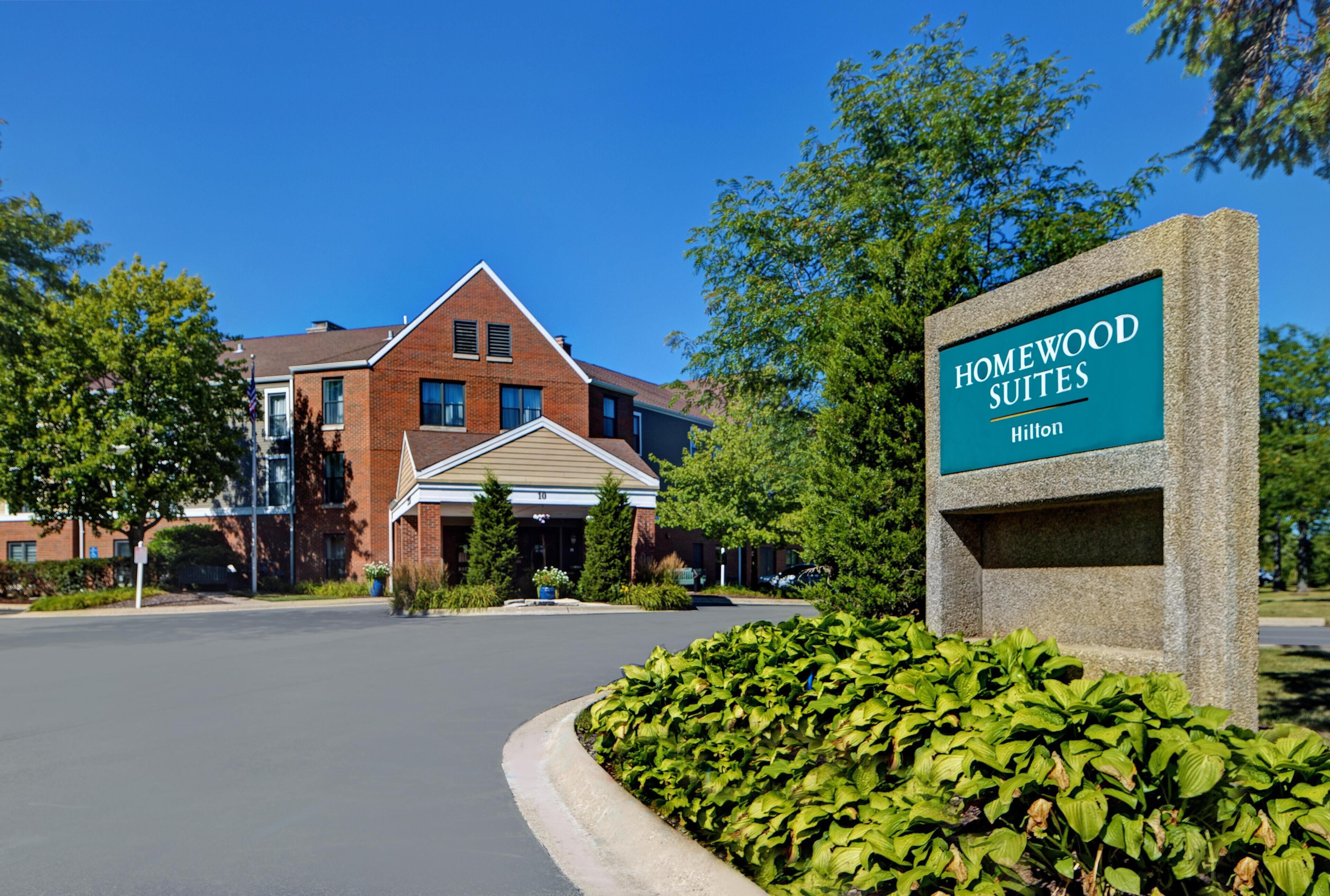 Entrada de un hotel con un letrero que dice 'Homewood Suites Hilton' y un edificio de ladrillo al fondo.