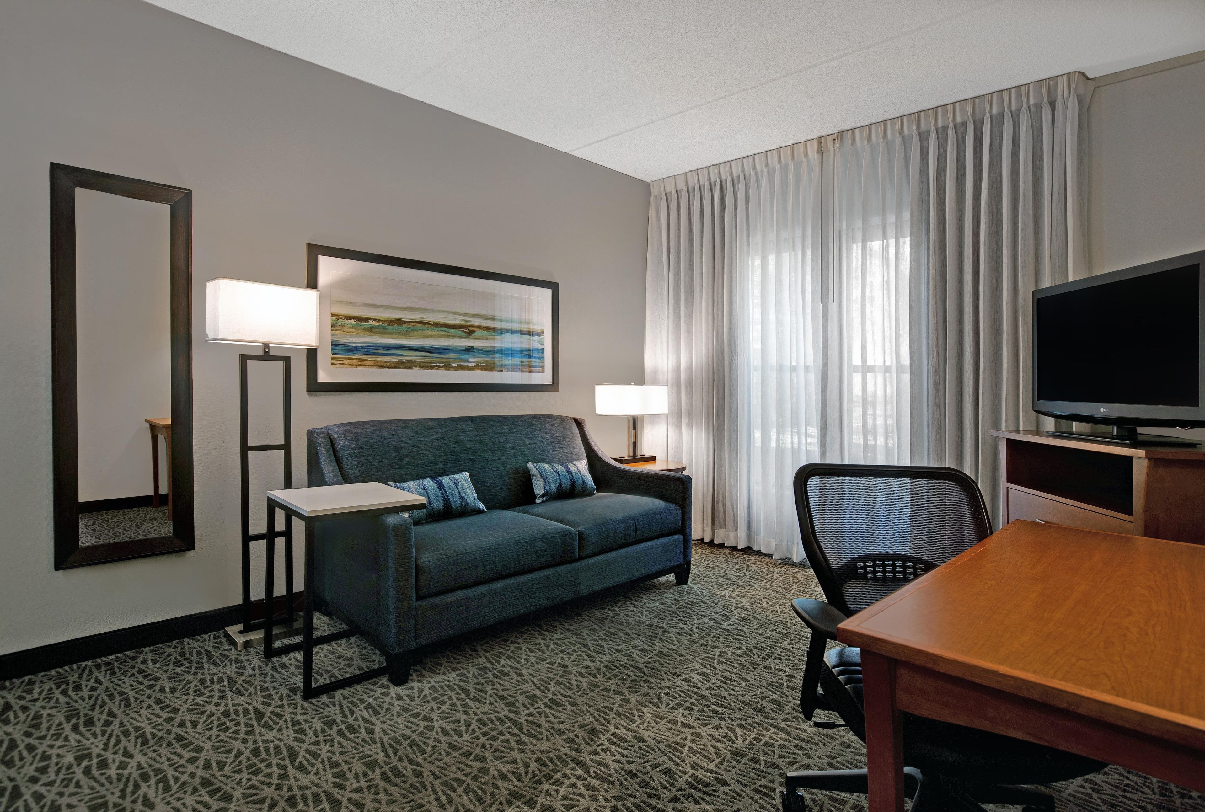 Quarto de hotel com sofá, televisão, escrivaninha, cadeira, abajur, quadro e janela com cortinas.