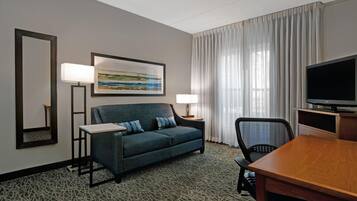 A hotel room with a sofa, a TV, a desk, a chair, a lamp, a painting, and a window with curtains.