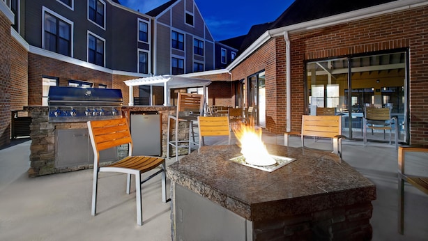 Een terras buiten met vuurkorf van steen, zitplaatsen en een barbecue.