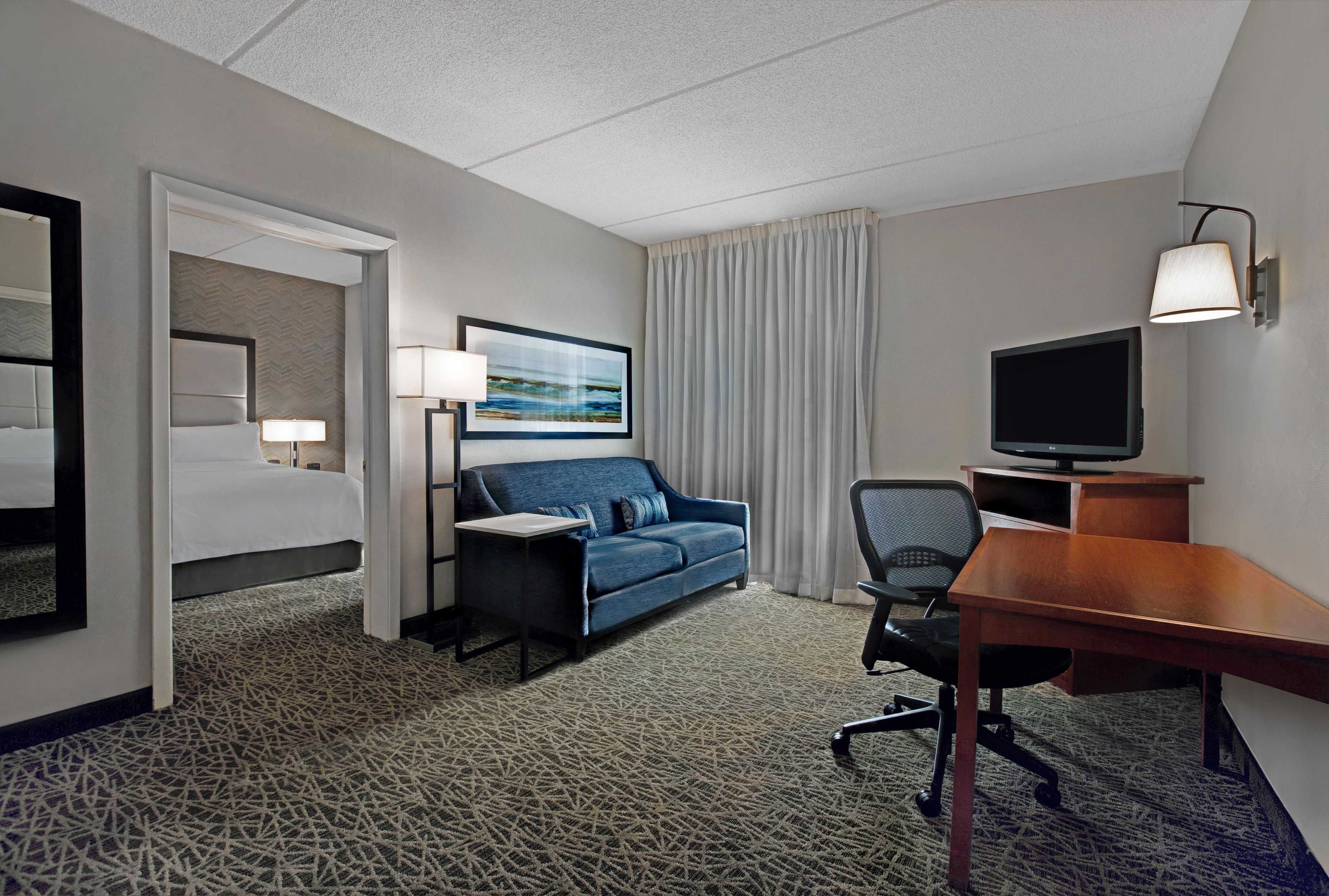 Una habitación de hotel con cama, sofá, escritorio con televisor y ventana con cortinas.
