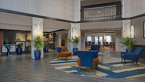 Een ruime lobby met een hoge plafond, stenen zuilen, en een centraal zitgedeelte met blauwe en bruine stoelen.