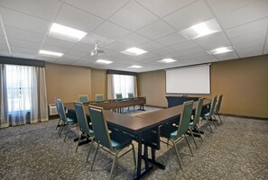 Ein Konferenzraum mit einem langen Tisch, Stühlen, einer Projektionsleinwand und einem großen Fenster mit Vorhängen.