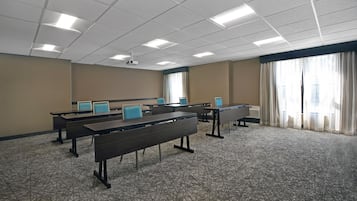 A conference room with multiple tables and chairs, a projector mounted on the ceiling, and large windows with curtains.