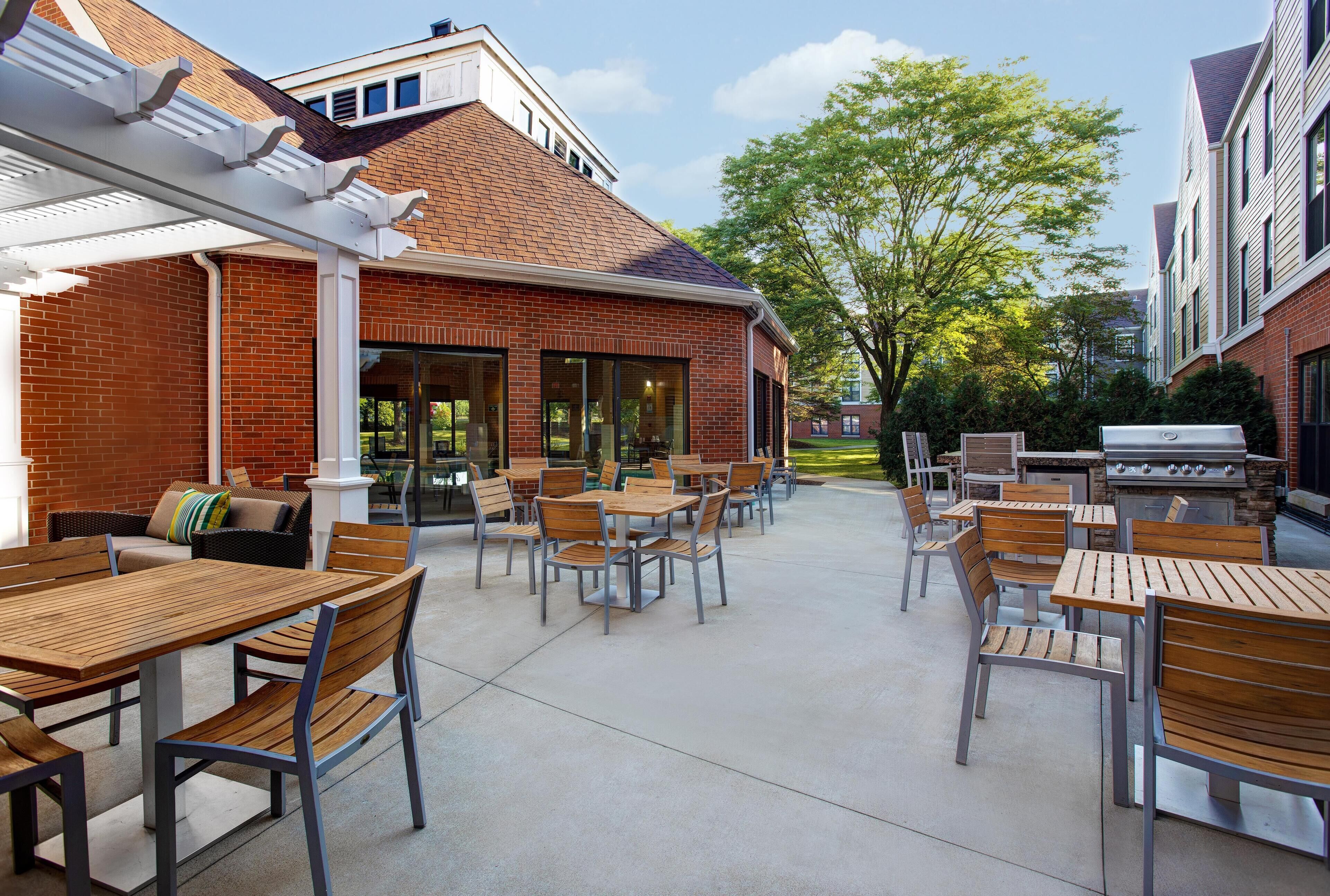 An outdoor patio area with wooden tables and chairs, a brick building, and a barbecue grill.