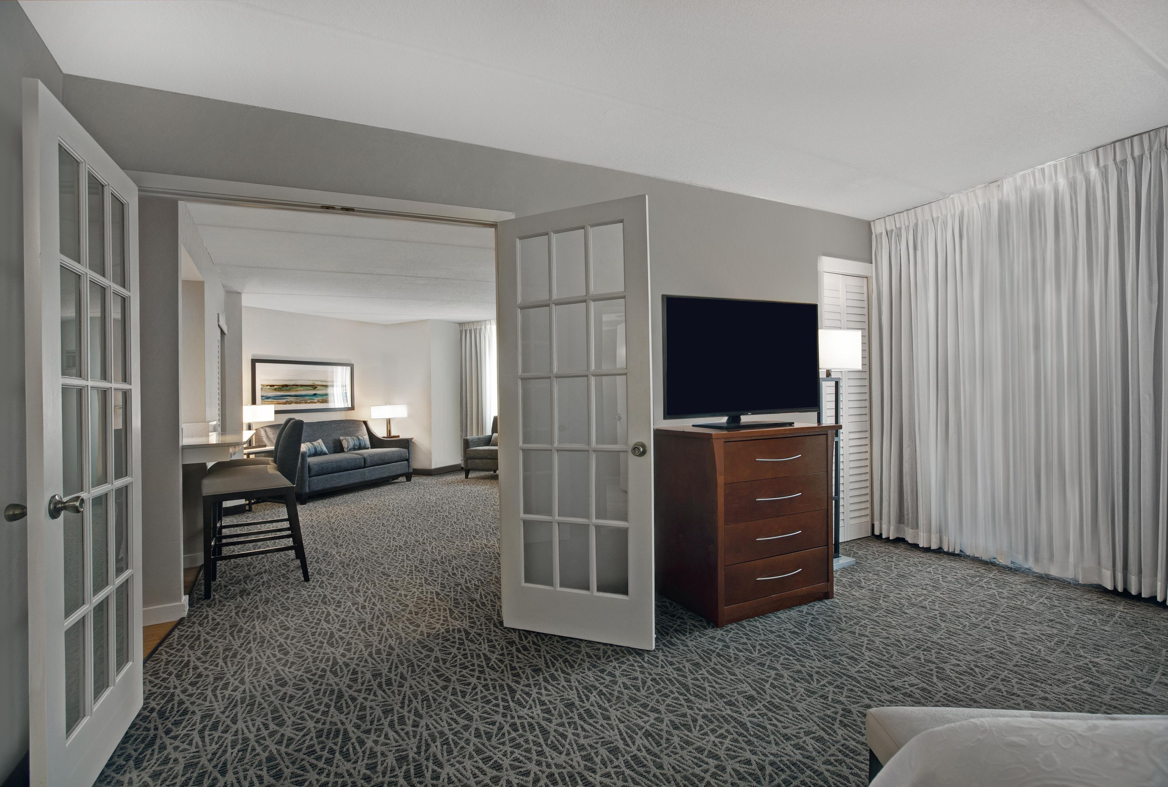 A hotel room with a television on a dresser, a sofa, a desk, and a large window with curtains.