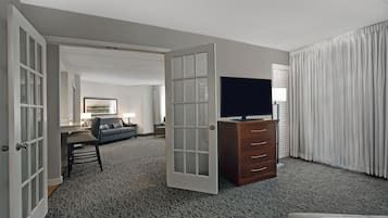A hotel room with a television on a dresser, a sofa, a desk, and a large window with curtains.