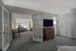 Habitación de hotel con televisor en una cómoda, sofá, escritorio y un ventanal con cortinas.