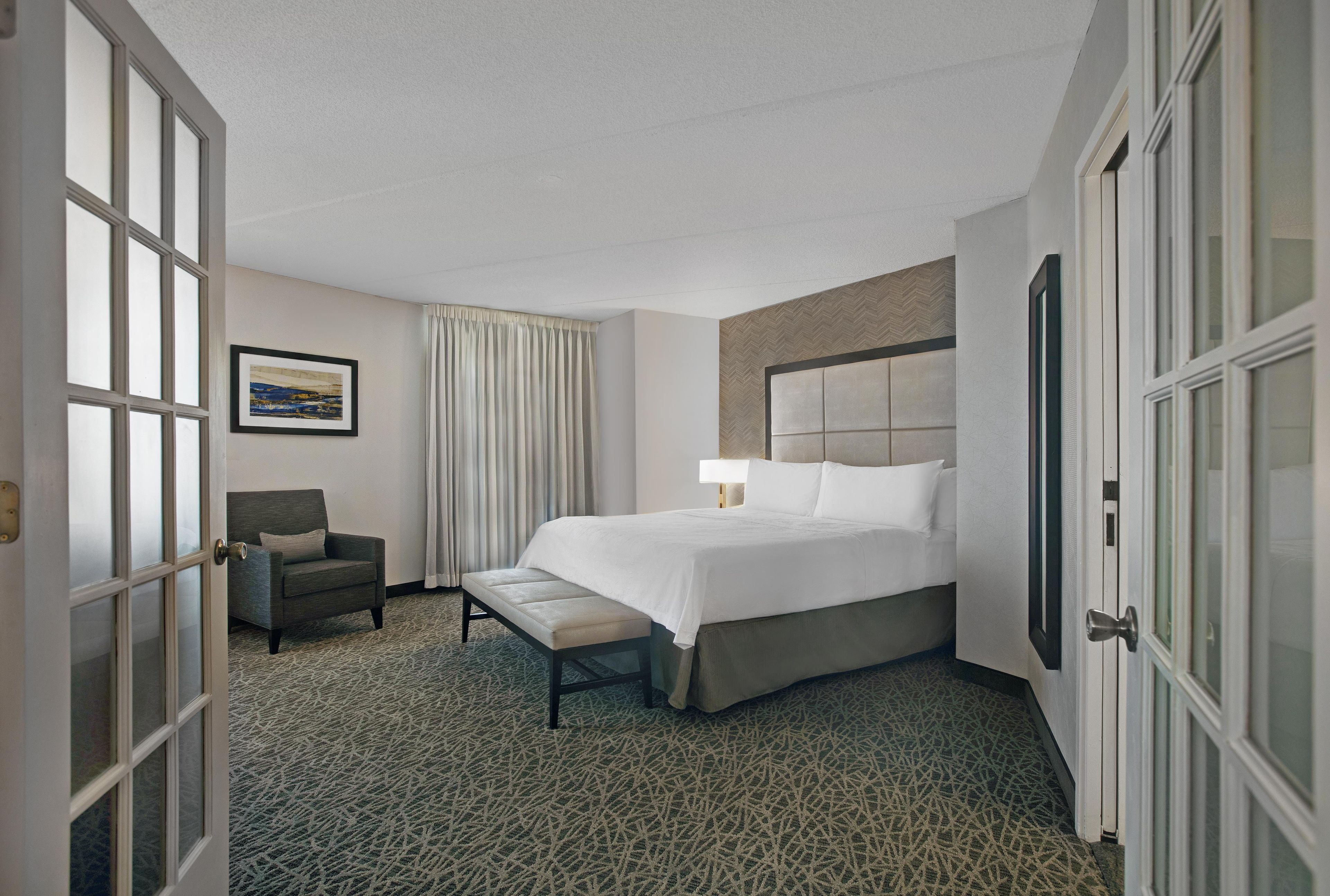 A hotel room with a bed, a chair, a bench, a picture on the wall, and a door.