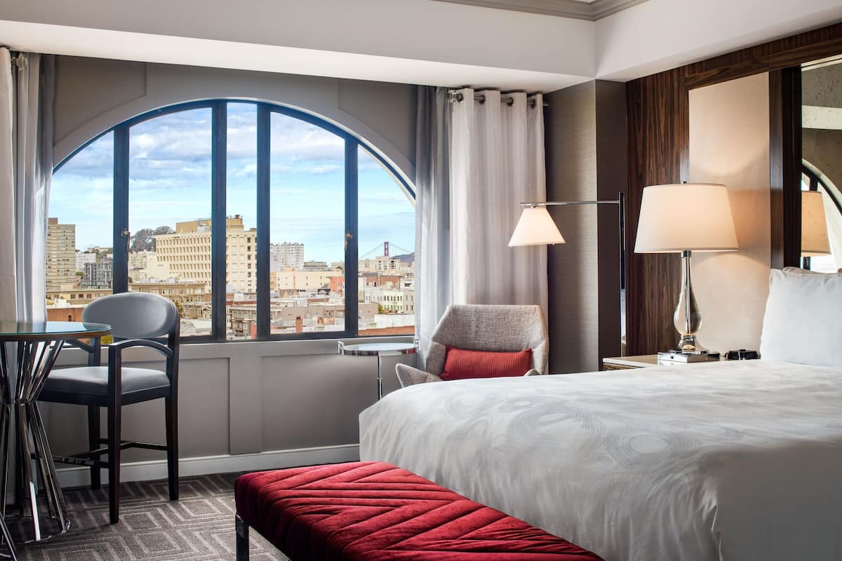 Habitación de hotel con un ventanal grande que ofrece vista a la ciudad, una cama con ropa blanca, un banco tapizado en rojo, una silla y una mesa.