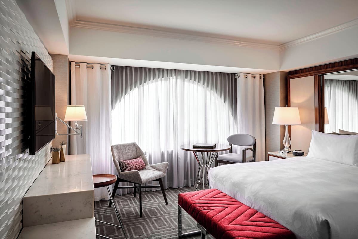 Habitación de hotel con una cama grande, un escritorio con lámpara, una silla, una mesita y vistas al exterior a través de cortinas translúcidas.