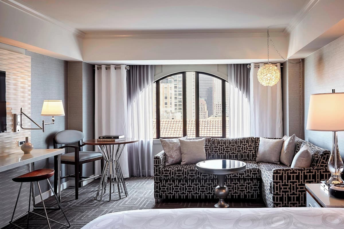 Una habitación de hotel moderna con un sofá estampado, una mesita redonda pequeña, un escritorio con una silla y un amplio ventanal con cortinas.
