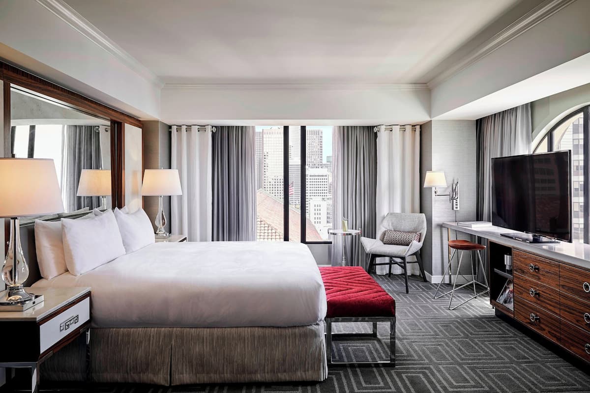 Habitación de hotel con una cama grande, televisión, escritorio con silla y vistas a la ciudad desde las ventanas.