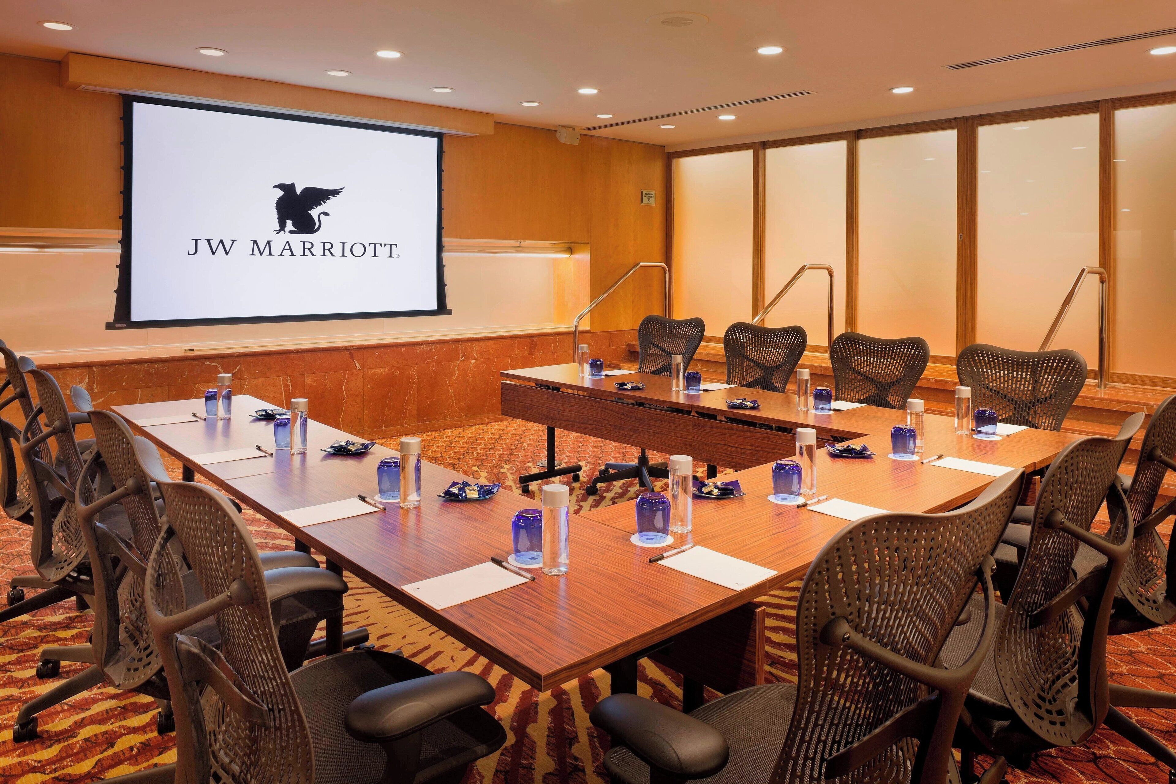 Un salón de conferencias con una mesa larga, sillas, una pantalla de proyector y un logotipo en la pared.