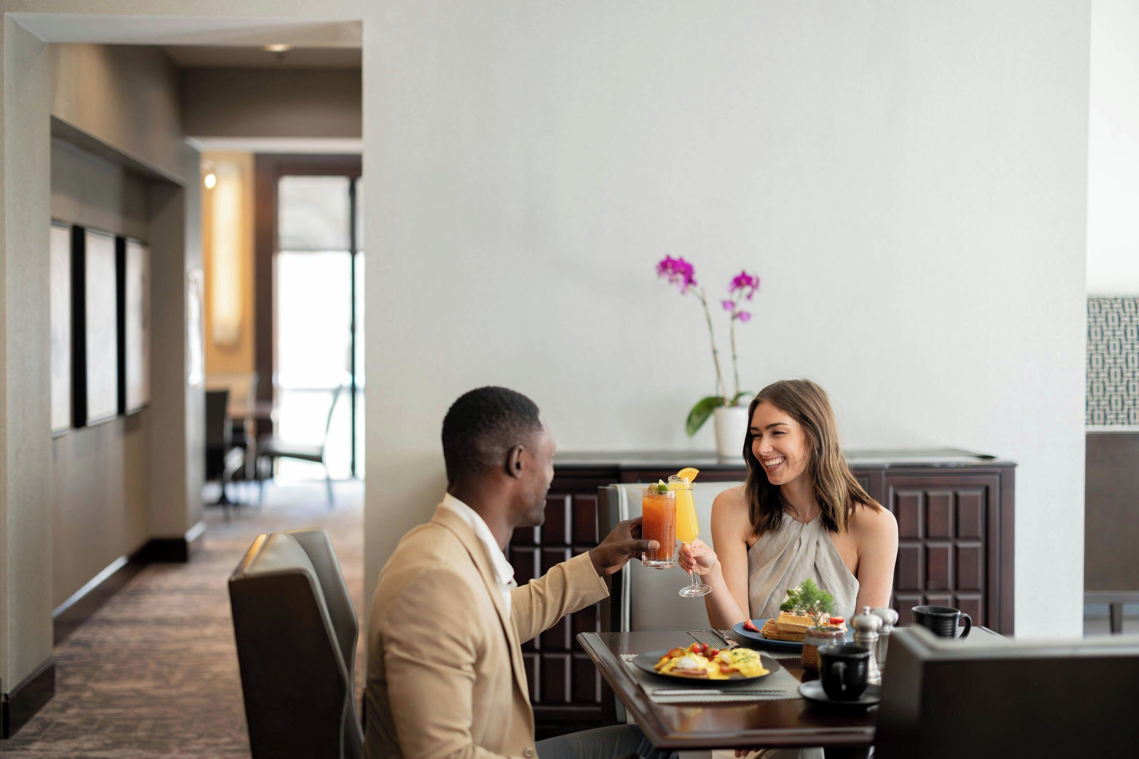 A man and a woman toasting with drinks at a hotel dining table, with a plant and a window in the background.