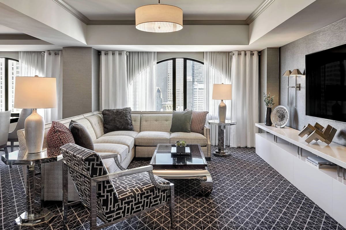 Una habitación de hotel moderna con un sofá modular, un sillón estampado, una mesa de centro de vidrio y un televisor de pantalla plana.