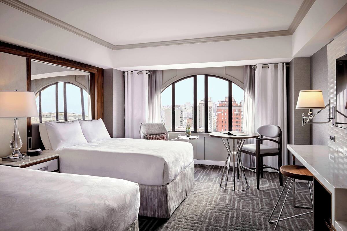 Habitación de hotel con dos camas, un escritorio, una silla, una mesita pequeña y un amplio ventanal con cortinas.