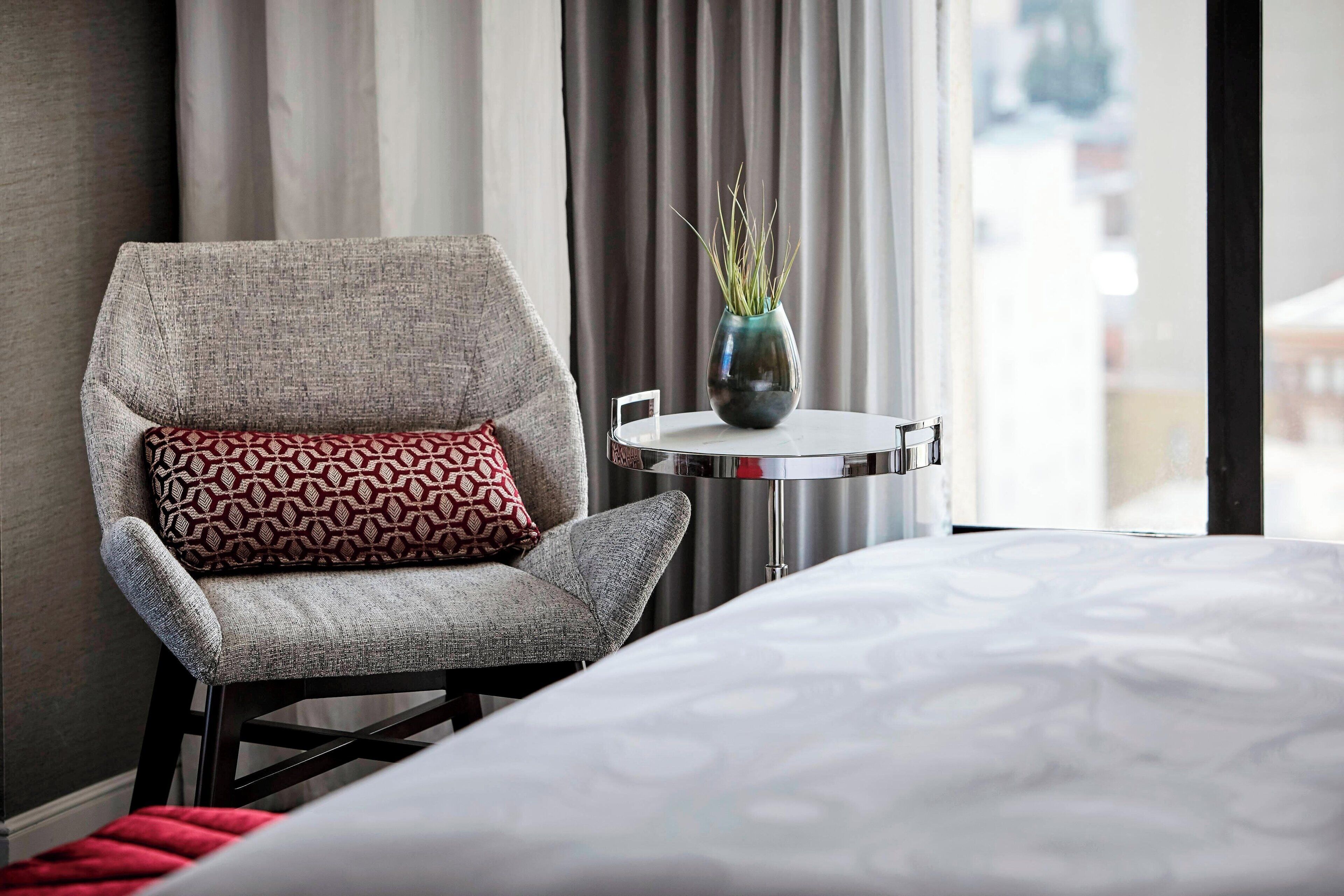Ett hotellrum med en grå fåtölj, en mönstrad kudde, ett litet bord med en vas och en säng med ett mönstrat överkast.